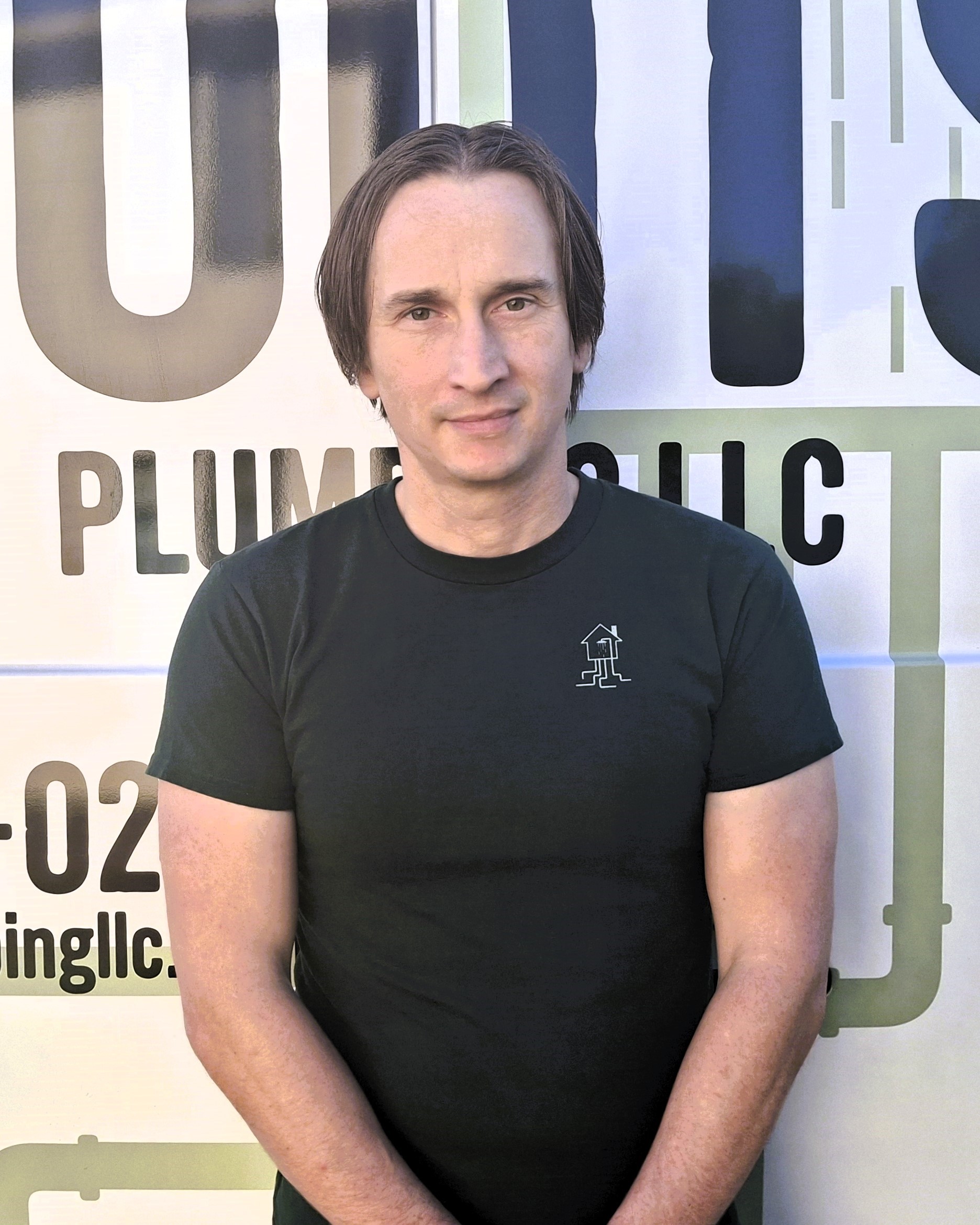A man with straight brown hair in a black t-shirt stands in front of a sign that reads PLUMB LLC with partial contact information visible.