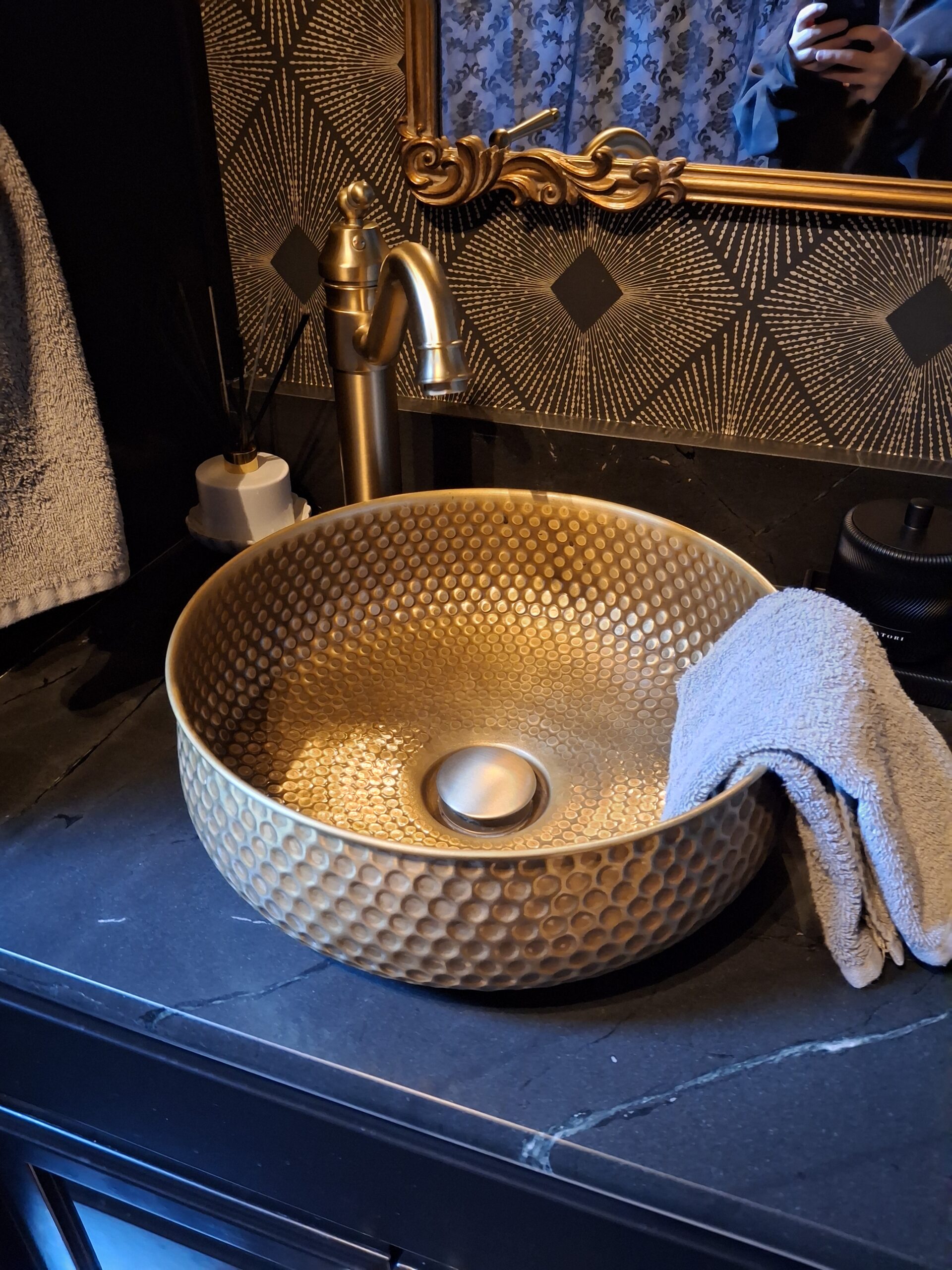 A gold textured vessel sink with a matching faucet, a gray hand towel draped over the edge, and patterned wallpaper in the background.
