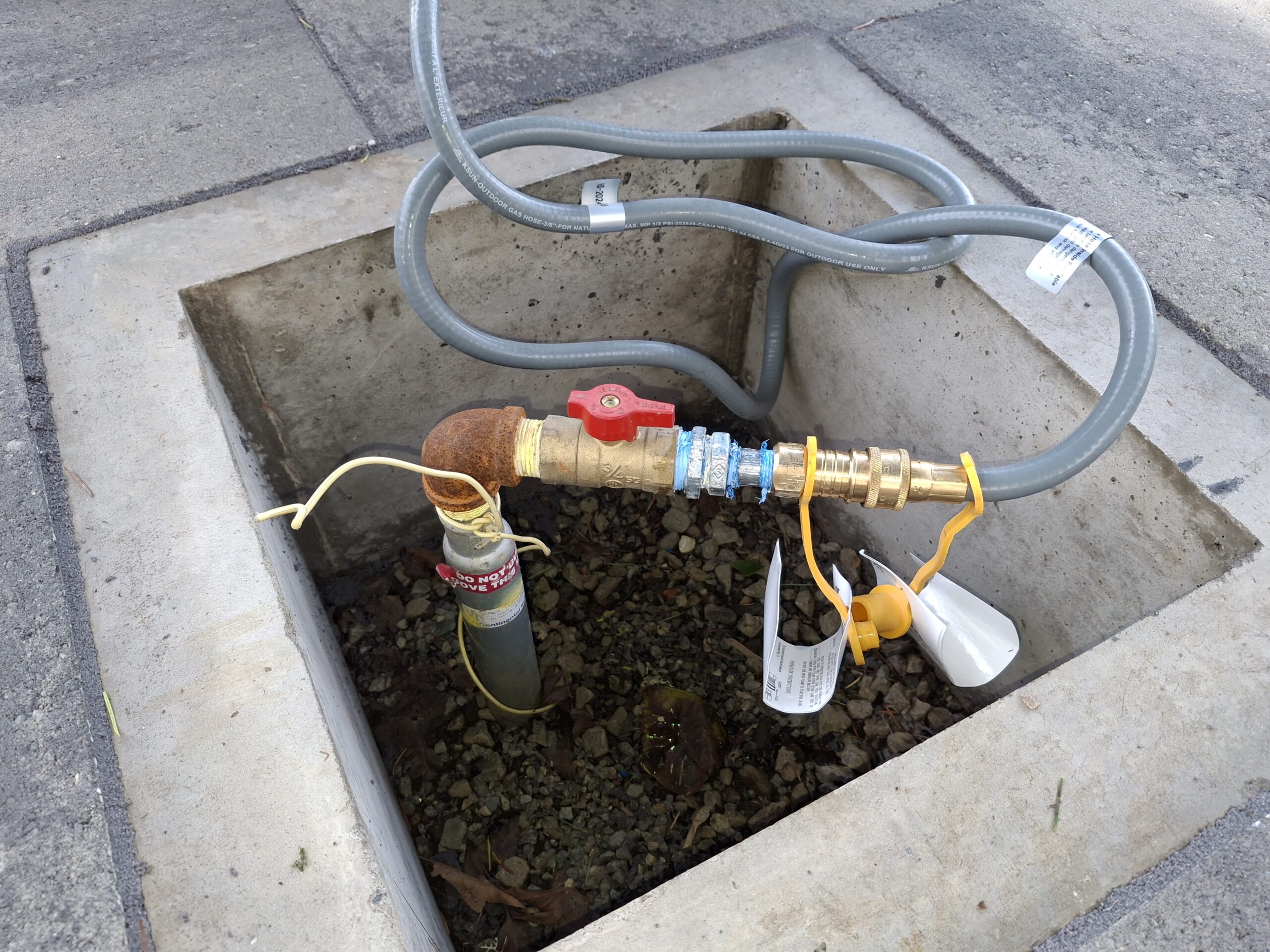 An exposed water pipe with a shut-off valve and attached hoses sits within a square concrete enclosure on a paved surface.