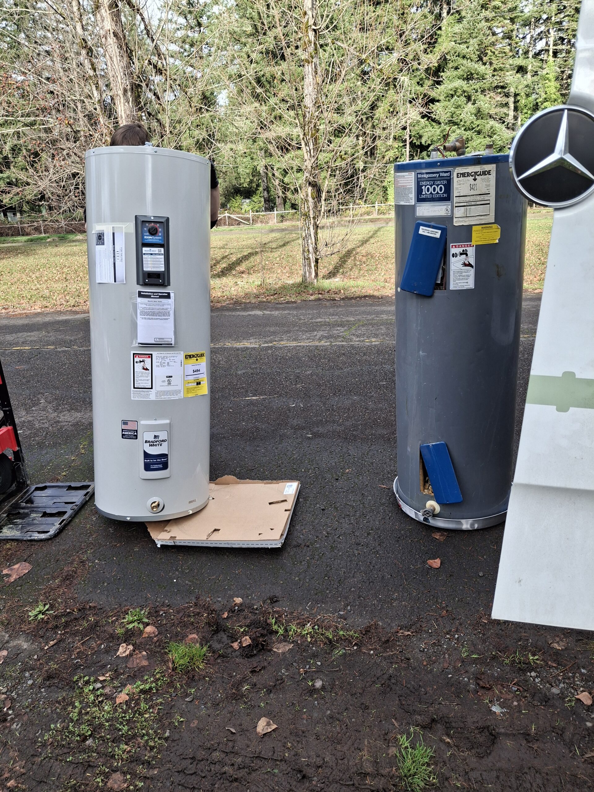 Two water heaters, one new and one old, are standing upright on an outdoor paved area near some trees. The new unit is on a cardboard sheet, and both have labels attached.