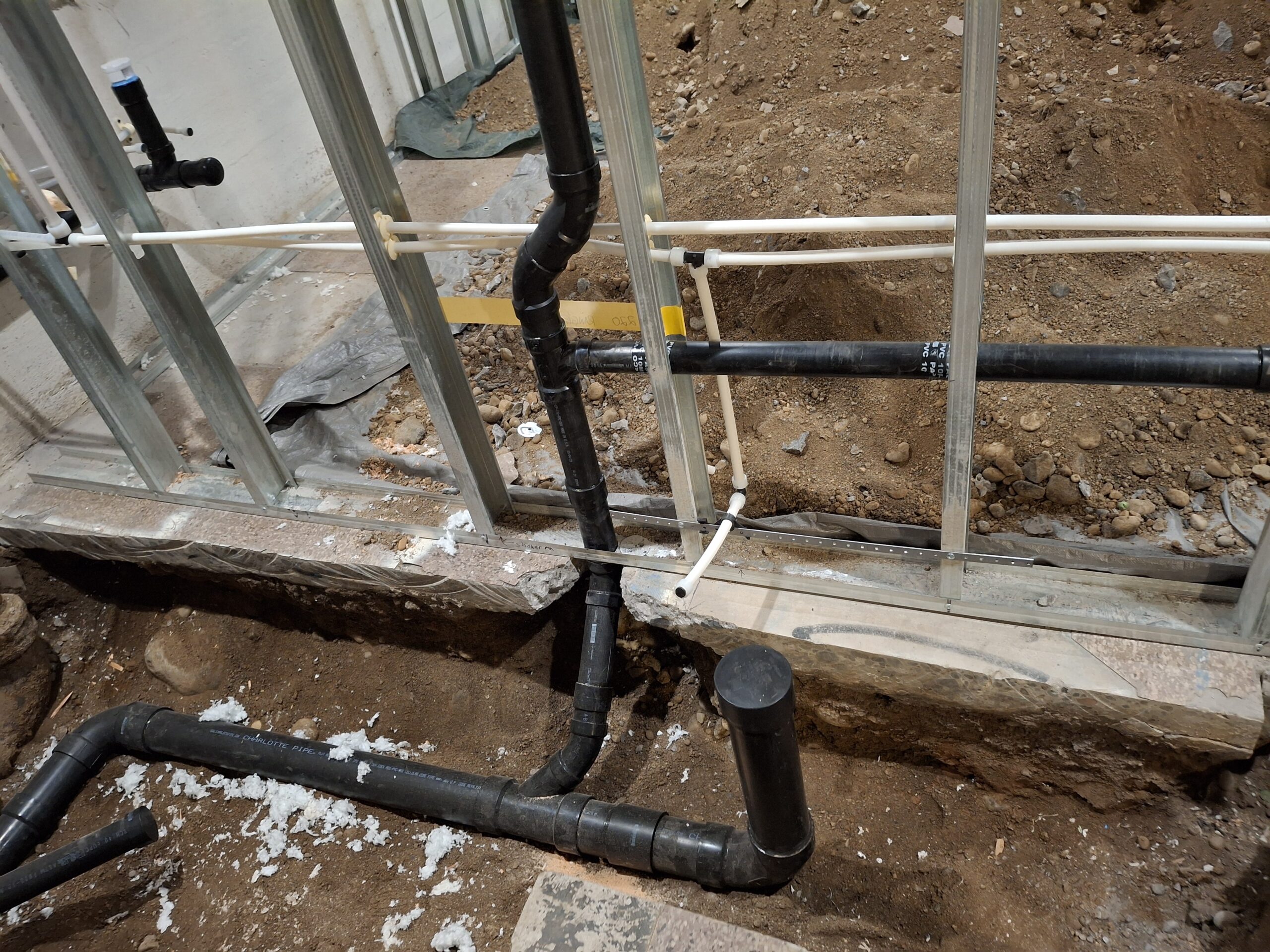 Plumbing pipes and PVC drainage system installed in a building under construction, with exposed soil and metal wall framing visible.