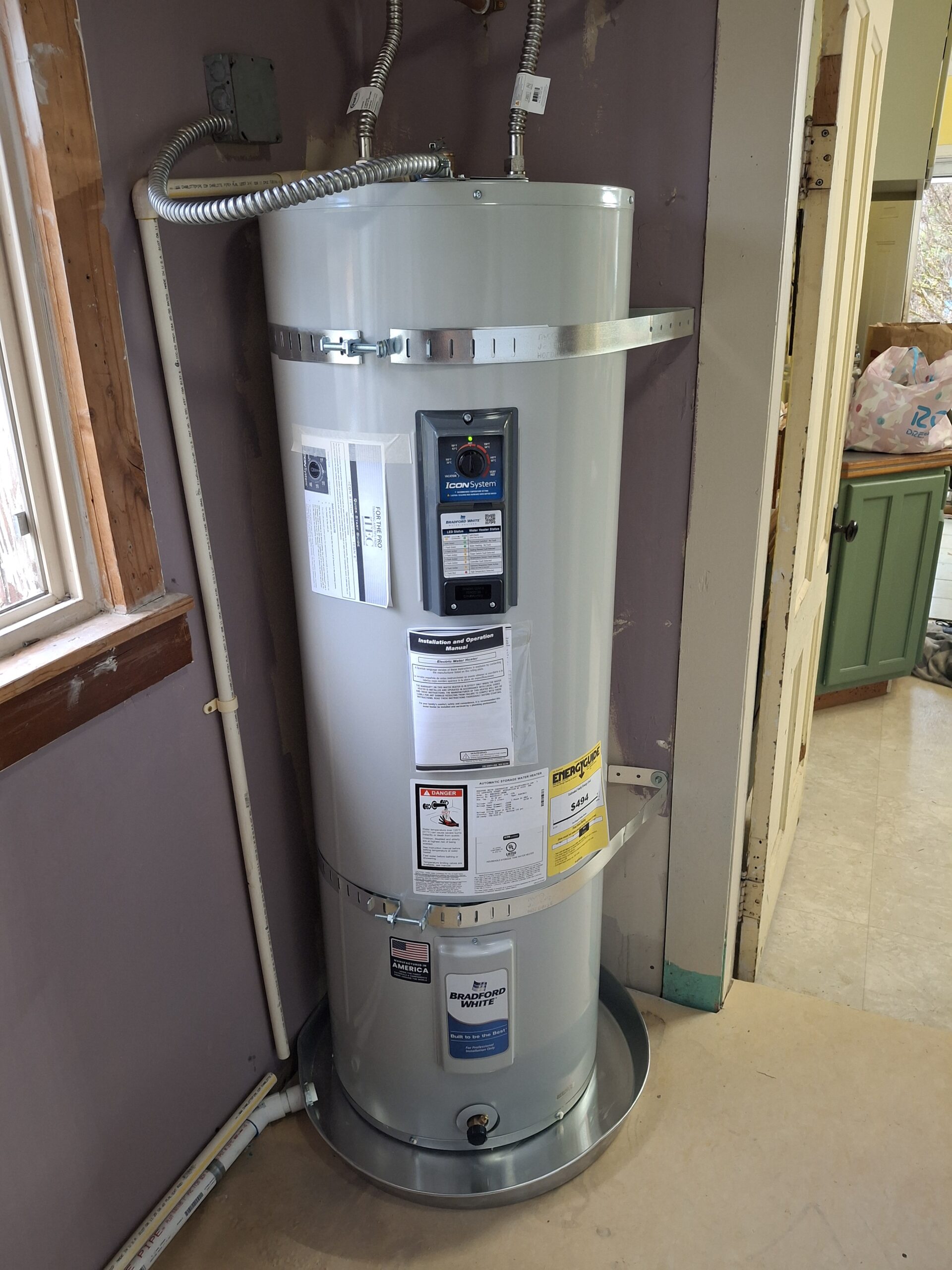 A new electric water heater installed in a corner, with attached labels, wires, pipes, and a metal drain pan underneath.